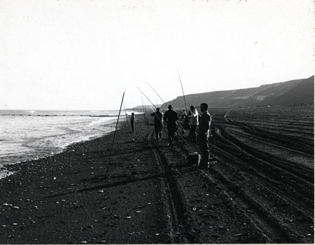 Photograph of seven men fishing on Horden beach; the photograph is taken looking south and shows the surface of the beach scored with tyre tracks; the men are standing with their rods on the beach looking out to sea; the photograph is dark and it would be difficult to identify the individuals