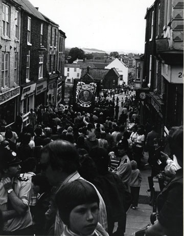 Photograph of the Horden Lodge banner being carried down Elvet Bridge in the City of Durham; the facades of the shops on Elvet Bridge and the buildings of Old Elvet can be seen; the banner which shows a portrait of Emmanuel Shinwell, a prominent Labour politician of the twentieth century