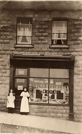 Photograph of the exterior of a shop showing the shop window and two windows above; outside the shop a woman and a girl are standing; the name M. Miller can be seen on a notice above the door and advertisement for Crawford's Biscuits can be seen in the shop window; the photograph has been identified as Miller's Cake Shop, Cotsford Lane, Horden, 1918-1920; the shop window is not identical with that identified as Miller's Cake shop in hord0059
