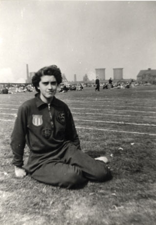 Photograph of a woman wearing a tracksuit, sitting on a sports field, with people in the distance and in the far distance the buildings of the colliery; she has been identified as D. Skidmore, Champion Runner, Horden, who apparently won the 100 and 200 Yards Race at Horden on 30 July 1955; the 100 Yards Race at Byker on 11 June 1955; the 100 Yards Race at Hebburn on 2 July 1955; 100 Yards Race at Gateshead on 27 August 1955