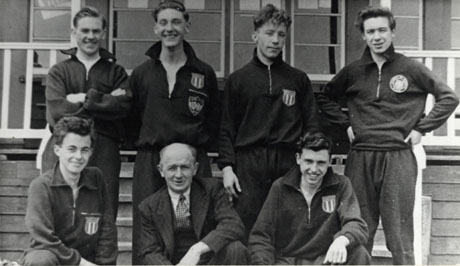 Photograph of six young men in tracksuits posed outside a wooden building with a middle-aged man in a suit; the photograph has been described as Horden Athletics Sports Club, 1955; the people have been identified as, left to right: Marshall; Watson; Kells; Hutchinson; Thompson; Hamilton(Trainer); Lee