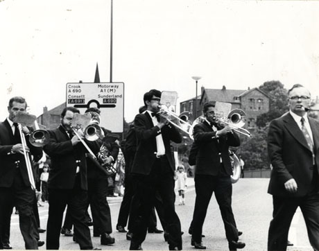 Photograph of nine members of the Horden Band playing their instruments and crossing a road in the City of Durham during the Miners' Gala; a man with a pipe can be seen in front of the band at the right of the photograph