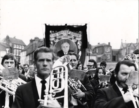 Photograph of six members of the Horden Band in the City of Durham playing their instruments in front of the Horden Lodge banner during the Miners' Gala; the faces of the players can be seen; buildings in the city can be seen indistinctly in the background; the banner showing the portrait of Harold Wilson can also be seen indistinctly