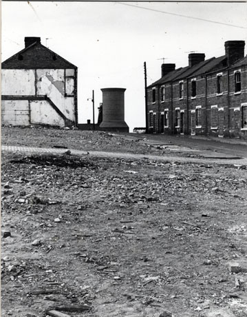 Photograph of the demolition of 2nd Street, Horden, showing a large open space with the end of a house, on the left-hand side of the photograph, in the distance; on the right of the photograph, a row of boarded-up terraced houses remains; at the end of the street a construction which may be a ventilation shaft may be seen