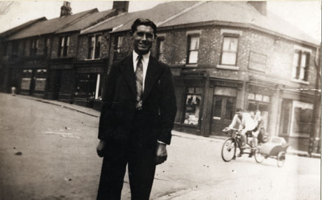 Photograph of Blackhills Road, Horden, showing, in the background of the photograph, the corner of one side of the road with shops along it; in the foreground a man, identified as W. Clark, is standing in the road dressed in a suit and tie; behind him, riding on a tandem bicycle with a sidecar, are a man and a woman, who have been identified as Mr. and Mrs. Peart