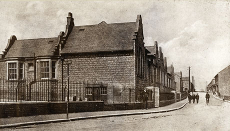 Photograph described as Horden Higher Tops Leading To Old 5th Street, showing the side and front of a school on the left of a street leading away from the camera; further unidentifiable buildings beyond the school and on the other side of the road can be seen; a man can be seen standing outside the railings surrounding the school and three men can be seen walking down the road towards the camera, and a fourth riding a bicycle away from the camera