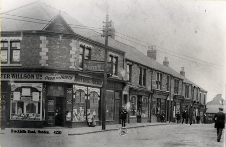 Postcard photograph entitled Blackhills Road, Horden. 4220, showing one side of the road with the facades of shops along its length; on the corner of the street the windows of a branch of Walter Willson Ltd., with the legends All Over The North and Money Saved On Every Purchase can be seen over the windows, in which the merchandise is indistinct; a bank and a shop belonging to G. W. Foster can also be seen; nine indistinct figures ca nbe seen on the road