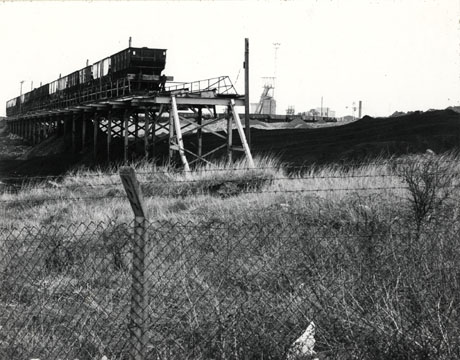 Photograph described as Teeming by Horden Colliery Facing South, 1976 showing the coal trucks on a rail built over coal waste; the winding gear of the colliery can be seen in the background to the right and a fence is in the immediate foreground of the photograph; the subject is the same as that in hord0112 and hord0199