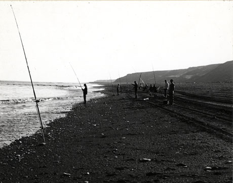 Photograph described as Angling Club, Horden, 1976, showing nine men fishing on the beach at Horden; the photograph is taken looking south and shows the figures of the men with the hills behind them and the surface of the beach scored with tyre tracks; the photograph, as in hord0041 and in hord0207, is dark, and the men would be difficult to identify