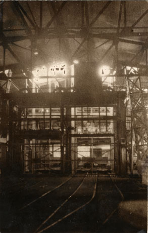 Photograph described as Hydraulic Decking Plant, Horden: The Cage, showing the interior of a large structure with steel beams in the roof and rail tracks on the floor; in the middle distance a large open steel structure which resembles two lift cages, approximately three quarters of the height of the building, can be seen