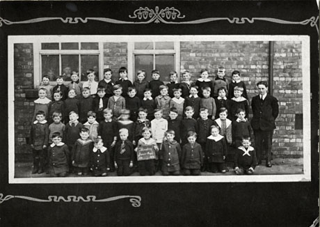 Photograph of a group of forty nine small boys, aged approximately seven years, posed in front of the wall of a brick building with a man, presumably their teacher; one of the boys on the front row is holding a notice reading: Horden Junior Boys 1919