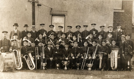 Photograph of thirty seven members of Horden Colliery Welfare Band, posed in font of the wall of a house; twenty seven of the men are in uniform and are carrying their instruments, and ten of the men are un suits; five trophy cups are on the floor in front of the men