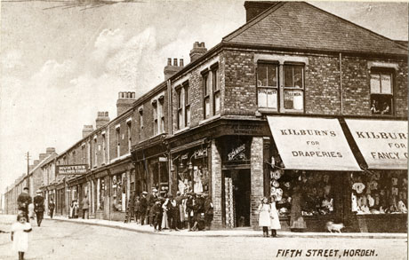 Postcard photograph entitled Fifth Street, Horden showing the corner and one side of a street receding from the camera; on the corner of the street is a shop with four windows and awnings on which is written Kilburn's For Draperies; a group of approximately eleven people are standing in front of two of the windows of the shop, and two girls, a woman and a dog in front of another; a sign reading Hunter's Wallpaper Stores can be seen further down the street; six adults can be seen further down the street and a child is standing in the road in the foreground