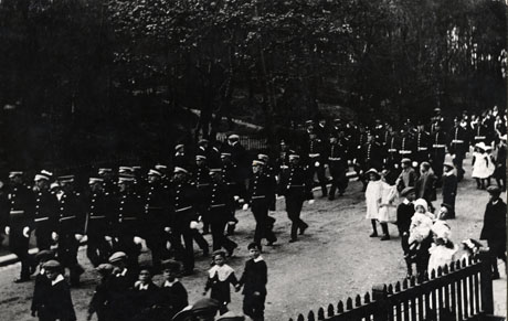 Photograph showing a procession of men in uniform moving across the picture from right to left; the procession is accompanied by approximately sixteen children running alongside the procession; a woman with two infants in her arms can also be seen; the men are marching past a heavily wooded area; the photograph has been identified as Procession to the Opening of St. Mary's Church, Horden