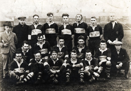 Photograph of the fifteen members of the Horden Rugby Team posed on a field with the backs of houses in the distance; with the members of the team are four other men