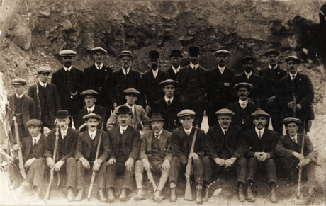 Photograph of a group of twenty five men posed against a wall of rock, wearing suites, ties, hats, and caps; the men are each holding a rifle; they have been identified as members of Horden Rifle Club in 1912