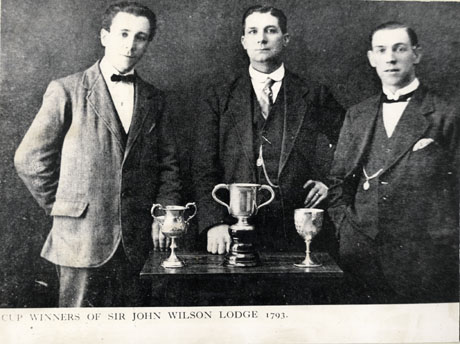 Photograph of three men, smartly dressed in suits and ties, standing behind a small table on which three trophy cups are standing; the photograph, which appears to be a photograph in a printed book or magazine, has the printed legend below it: Cup Winners of Sir John Wilson Lodge 1793