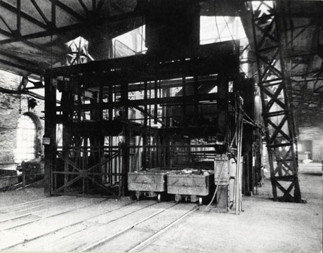 Photograph of the cage at the Hydraulic Decking Plant at Horden Colliery, as in 0236; the photograph, as does 0236, shows the interior of a large building with steel girders in its roof and rails on its floor; in the foreground the cage can be seen with two trucks standing in one side of it