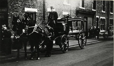 Photograph of a horse-drawn hearse standing outside a terrace of houses; the hearse is drawn by two horses and a man is sitting on the hearse, which does not contain a coffin; further along the pavement an indistinct group of four people can be seen; at the right- hand edge of the photograph the bonnet of a large car can be seen; the photograph has been identified as Tommy Gear's Funeral, Horden, 1960 (Fruit and Vegetable Wholesaler)