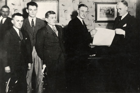 Photograph of six men dressed in suits and ties standing in front of an interior wall decorated with a patterned wallpaper and two pictures; one man is being handed a piece of paper by another and the others are looking on; the photograph has been described as Charlie Turnbull Receiving Certificate of Merit, Horden