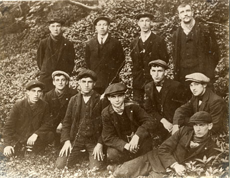 Photograph of a group of eleven young men in suits and caps posed against trees and bushes; the photograph has been described as depicting a Group of Miners, Dene Holme, Horden, 1926