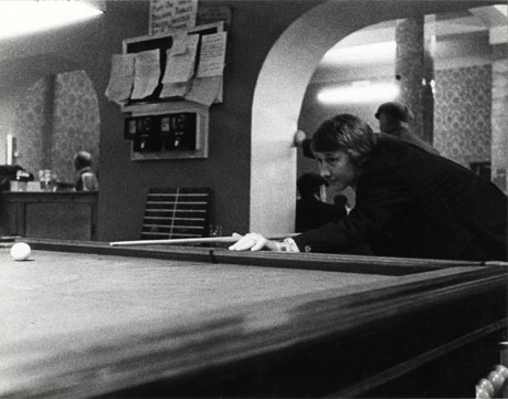 Photograph showing, at the front of the picture, the surface of a snooker table and a young man with a snooker cue in his hand leaning on the table preparing to play; behind the snooker table, a wall with two arches cut in it can be seen; beyond one of the arches a bar with a man behind it can also be seen; the photograph has been identified as the Horden Big Club