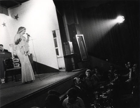 Photograph of a woman in an evening dress standing on a stage holding a microphone; behind her a musician can be seen indistinctly and in front of her an audience of people sitting at tables drinking can be seen; the photograph has been identified as Horden Big Club