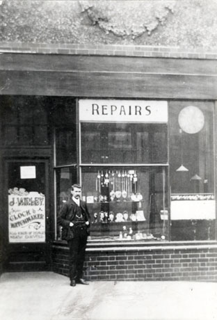 Photograph of the exterior of the shop of J. Varley, Clock and Watchmaker, showing the contents of the shop window and a man, presumably Mr. Varley, standing in front of the window; above the shop a decorative wreath on the wall can be seen, indicating that the shop is part of a large decorative building such as a cinema