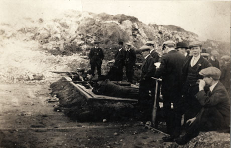 Photograph of four men lying on a raised platform sideways to the camera about to fire rifles at a target out of the picture; behind the men shooting are approximately thirteen men observing the proceedings; behind all the men rough hillside can be seen; the men have been identified as members of Horden Rifle Club