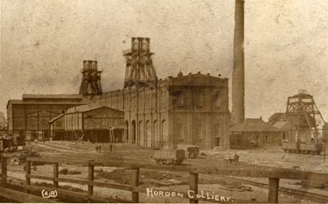 Postcard photograph entitled Horden Colliery, described as Horden Colliery, showing North, South, and East Shafts; the photograph shows the surface building of the colliery with the top of the winding gear behind, a chimney to the right of the building and rails and trucks in the front of the building
