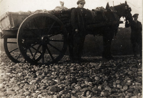 Photograph of a cart, drawn by a horse, standing on a stony surface, sideways to the camera; a man is standing in front of the cart and another is standing at the horse's head; the words, J. King, can be seen on the side of the cart; the photograph has been identified as Jim King(Contractor) Stone Loading From Beach (Hardcore For Coast Road, 1924)