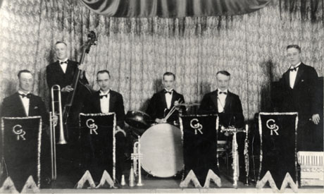 Photograph six men in evening dress on a stage with musical instruments; behind the men there is a curtain and in front of them stands with the initials G R; they have been identified as Gilbert Ridley's Band about 1935