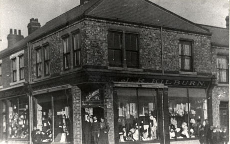 Photograph of the exterior of a shop on the corner of a road; four windows of the shop can be seen with clothing in them but the contents of the windows cannot be seen in detail; above the windows are the words:Horden Colliery Post Office J. R. Kilburn, and above the doorway are the words:Bon Marche; two men and a woman can be seen standing in the doorway of the shop