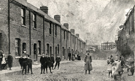 Postcard photograph entitled Sea View Horden, showing the left-hand side of a street of terraced houses running away from the camera; at the end of the street surface buildings of the colliery can be seen, possibly in the process of being built; the surface of the road is rough stones; fifteen children can be seen standing in the road and sitting on the pavement; ten adults can be seen standing on the pavement and in doorways; it has been suggested that Sea View later became Second Street