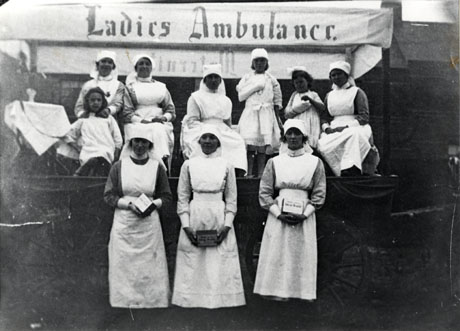Photograph of four women dressed in nurses' uniform sitting on a cart with three small girls, two of whom have a bandaged arm; three other women in nurses' uniform are standing in front of the cart; behind the women on the cart is a banner bearing the words Ladies Ambulance; the three women on the ground are holding collecting boxes, on which there are illegible words, one of which may be hospital