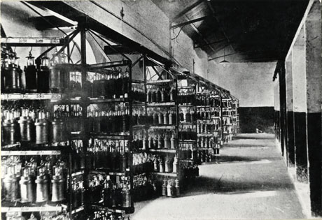 Photograph of the interior of a large building with what appear to be pillars down the right-hand side of the picture; on the left-hand side is metal racking from which electric miners' lamps are suspended in rows; above each lamp a number can be seen; behind the racking the top of large windows can be seen