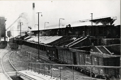 Photograph showing, on the left of the picture, part of an indistinct building in the distance; beside this building, a rail track, on which there is a truck, winds into the distance;beside the track, are further tracks, in the middle of the picture, on which three trucks with the initials N E can be seen; on the right side of the picture, a large structure, identified as Kopper's Coke Ovens, stretching the length of picture, can be seen