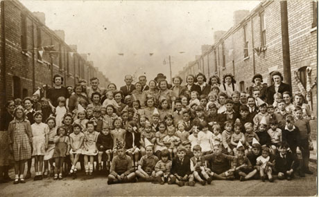 Photograph of approximately a hundred men, women, and children grouped across a street; the tops of the terraced houses on either side of the group can be seen; above the group a line of bunting can be seen; the occasion for the photograph has been identified as a street party in celebration of either Victory in Europe Day( 8 May 1945) or Victory in Japan Day ( 15 August 1945 )