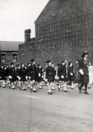 Photograph of a parade of Girl Guides being led by a Guider along a road with the blank wall of the end of a house behind them; at right angles to the wall the facades of terraced houses can be seen in the distance; indistinct figures watching the parade can be seen behind the Guides; the photograph has been described as Sunderland Road, Horden Cowell Street Procession Girl Guide Brigade