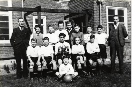Photograph of thirteen boys and two men posed in front of a brick building; eleven of the boys are wearing football strip and one boy is holding a trophy cup; the photograph has been identified as portraying the football team of Horden Modern School
