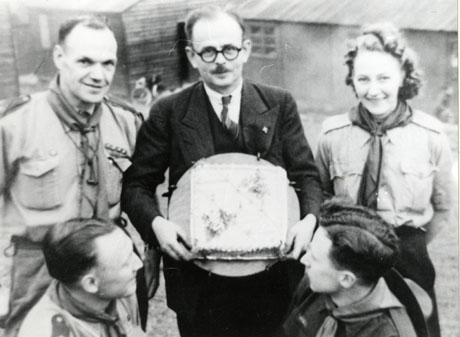 Photograph showing a front view of a middle-aged man, holding an unidentifiable object, flanked by a man in Scout uniform and a woman in Scout uniform; at the front of the picture two men wearing Scout uniform can be seen in profile; behind the group two huts can be seen indistinctly; the photograph has been identified as marking the First Birthday of the Scouts' H. Q. in Horden