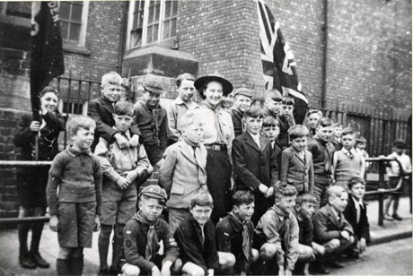 Photograph of twenty five boys posed on the pavement with a woman in Scout uniform outside a large brick building, possibly a school; six of the boys are wearing the uniform of the Cub Scouts; one boy is holding a Scout flag, and a Union Jack is being held by a boy, at the back of the group, who cannot be seen; the photograph has been identified thus: A Pack Cubs Victory Parade, May 1945