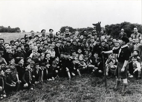 Photograph of a group of approximately fifty boys, aged between approximately seven and ten years, posed in the open air; most of the boys are wearing the uniform of the Cub Scouts; eleven adults, including a clergyman and Scouter, can be seen behind the boys; in front of the boys a Scouter is standing holding a pole with a wolf's head on top; the photograph has been identified thus: Easington and Castle Eden District Wolf Cub Rally Held at Horden, 1952. (Haswell and Horden Pack of Cubs)
