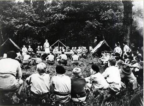 Photograph showing the back of a circle of Scouts looking towards three tents set up against a background of large trees; in front of the tents a trestle table has been erected and fifteen adults, including a clergyman and a Scouter, can be seen sitting behind it; in front of the table a figure in Scout uniform is standing addressing the group; the photograph has been identified as showing Lord Barnard officially opening the camp site Easington and Castle Eden Boy Scout Association