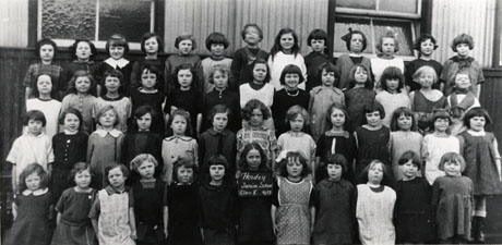 Photograph of a group of fifty one girls aged, approximately ten years, posed outside a corrugated iron building; a girl on the front row is holding a notice which reads: Horden Junior School Class 8 1927
