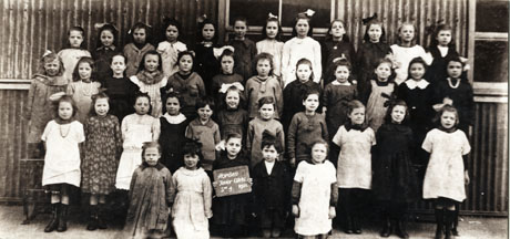 Photograph of a group of girls, aged approximately nine years, posed outside a corrugated iron building; a girl on the front row is holding a notice which reads: Horden Junior Girls Standard 4 1921