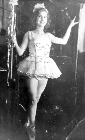 Photograph of a young woman wearing a tutu and a tiara, holding a wand with a star on its tip, and standing on pointes; she appears to be resting her left hand on an opened door and there appears to be a brick wall to her right; she has been identified as Singing Artiste, Horden