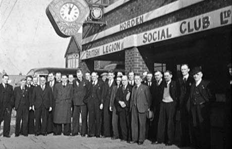 Photograph of twenty six men in suits standing posed outside a building on which the following words can be seen: Horden British Legion Social Club Ltd.; a clock above them on the building has the word Remembrance on it; behind the men the top of a single-decker bus can be seen
