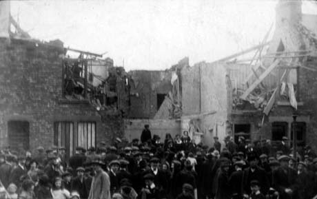 Photograph showing the ruined walls of a building with the interior of a room and the first floor of the house next to it exposed and the side of the chimney stack of the house next to that; on the other side from the exposed rooms, the front of a building has blocked up windows; crowds of people are standing in front of the ruins; the photograph ahs been described as Big Club After The Fire, Horden