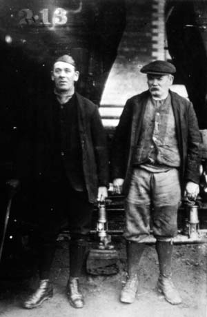 Photograph showing one man at the left of the photograph wearing a leather cap and carrying a miner's lamp; the photograph is too dark for the rest of his clothing to be seen; accompanying him is a man wearing a jacket, waistcoat, breeches, gaiters, boots, and a cap; he is also carrying a miner's lamp; they have been described as Two Miners, Horden